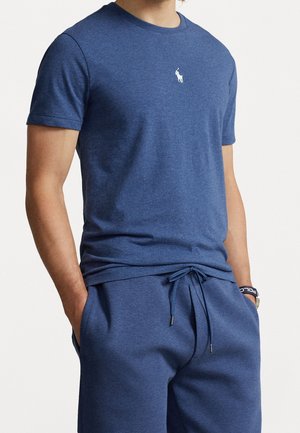 Mann trägt blaues T-Shirt mit kurzen Ärmeln und passende blaue Shorts mit Kordelzug, Hände in den Taschen, vor einfarbigem Hintergrund.