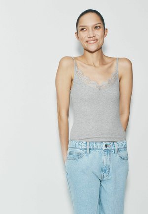 Smilende kvinde iført en grå camisole med blonder og lyseblå denimjeans, stående med hænderne bag ryggen på en ensfarvet baggrund.