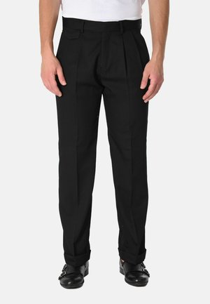 Uomo che indossa pantaloni neri plissettati con orli risvoltati, abbinati a scarpe monk strap in pelle nera e una camicia bianca infilata dentro.