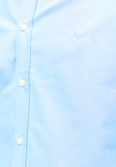 Ανοιχτό μπλε πουκάμισο με κουμπιά και μικρό κεντητό λογότυπο αλεπούς στο αριστερό μέρος του στήθους, με μαλακή υφή υφάσματος που φαίνεται σε κοντινή λήψη.