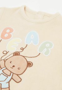 Beiger Baumwoll-Body mit einem gestickten Cartoon-Bären, der bunte Ballons hält. Mit rundem Ausschnitt und Druckknopfverschluss im Rücken.