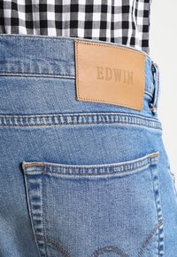 Light blue denim jeans feature a leather patch embossed with "EDWIN" at the back, with detailed stitching and a fifth pocket.