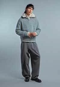 Jonge persoon die een grijze gebreide trui met een lichte kraag draagt, losse grijze broek en zwarte schoenen, staand tegen een effen grijze achtergrond.