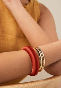 Röd satinsmycke bredvid två metallarmband—ett slätt guld och ett hamrat guld—på en bar handled mot en ljus bakgrund.