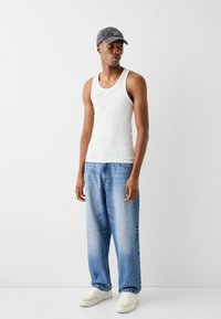 Top bianco senza maniche, jeans larghi azzurri chiari, sneakers bianche e un cappellino grigio con testo. L'outfit ha un aspetto casual e rilassato.