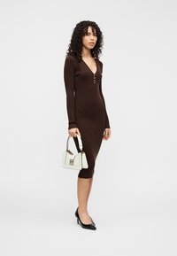 Καφέ πλεκτό φόρεμα με ριμπ ύφανση, V-λαιμόκοψη και λεπτομέρειες με κουμπιά, συνδυασμένο με λευκή τσάντα και μαύρες γόβες με μυτερή μύτη.