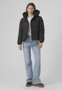Zwarte pufferjack met capuchon, doorgestikte ontwerp, voorzijde met rits en elastische manchetten. Gecombineerd met lichtblauwe flare jeans en stevige zwarte schoenen.
