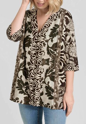 Donna che indossa una camicetta larga con motivi astratti e foglie in marrone, nero e beige, abbinata a jeans azzurri.