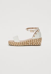 Sandales compensées blanches en cuir avec une plateforme en jute tressé, une sangle à la cheville avec boucle et des clous dorés sur le dessus.