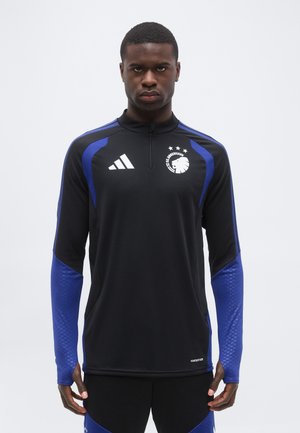 Mężczyzna ubrany w czarno-niebieską sportową bluzę z długim rękawem z logo FC Kopenhaga i marką Adidas stojący na jednolitym tle.