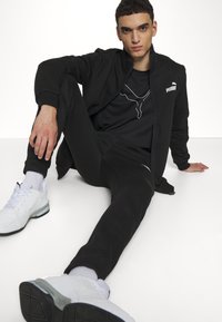 Black athletic tracksuit with zip-up jacket, tapered pants, and a black t-shirt featuring a white pattern. Shoes are white with gray accents.