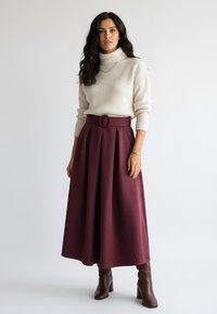 Kremowy prążkowany sweter z golfem w połączeniu z bordową spódnicą o kroju A, ozdobioną paskiem z okrągłą klamrą. Brązowe, sięgające kolan botki na obcasie.