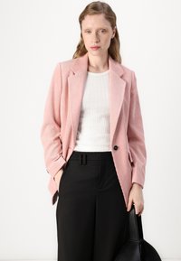 Blazer en velours côtelé rose avec revers, fermeture à un bouton, et un haut texturé blanc. Pantalons noirs à coupe droite et sac.