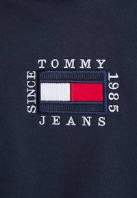 Marinblått tyg med en broderad rektangulär logotyp med röda, vita och blå ränder, samt vit text som lyder "SINCE TOMMY JEANS 1985."