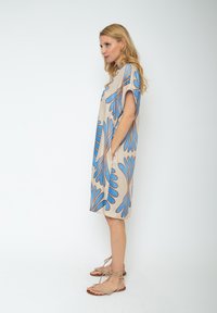 Beżowa sukienka z krótkim rękawem, ozdobiona niebieskimi i brązowymi wzorami kwiatowymi, z bocznymi kieszeniami i luźnym krojem. W zestawie z beżowymi sandałami na ramiączkach.