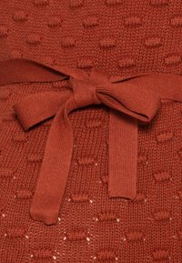 Kopparfärgad stickad tyg med texturerade prickar och ett knytbälte. Varpen har små öppna hål för en ventilerande design.