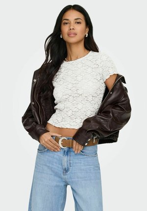 Donna con capelli lunghi e scuri indossa un top corto in pizzo bianco, una giacca di pelle marrone scivolata sulle spalle, jeans azzurri e una cintura marrone con fibbia argentata.