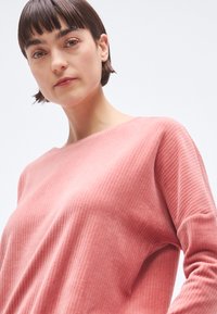 Chemise rose côtelée à manches longues avec un large décolleté et une texture douce. Vue en gros plan mettant en évidence les détails du tissu et la coupe.