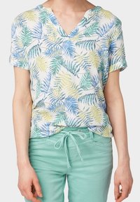 Camisa de manga corta y cuello en V en blanco con patrones de hojas verdes y azules. Combinada con pantalones de chándal verdes claros con cordón. Textura de tela suave.