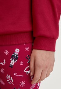 Rode sweatshirt met geribbelde mouwen, gecombineerd met gebloemde roze leggins met ontwerpen van een notenkraker, sneeuwvlokken, hulst en zuurstokken.