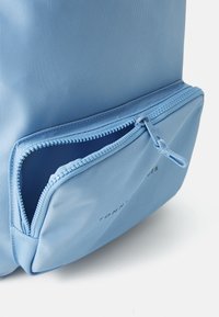 Sac à dos en nylon bleu clair avec une poche avant zippée et une surface texturée. Logo "TOMMY HILFIGER" visible sur le côté.