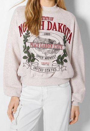 Bluza z czerwonym napisem „North Dakota”, mapą USA, „Peace Garden State” i ozdobnymi zielonymi liśćmi, noszona z białymi spodniami.