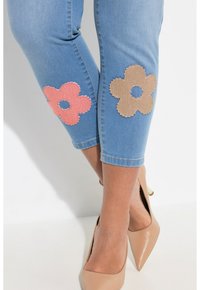 Pantalones capri de mezclilla con grandes apliques de flores rosas y beige en las rodillas. Tejido azul claro con una textura suave y un diseño ajustado.