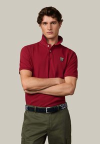 Camisa polo roja con cuello doblado, mangas cortas y un logo en el lado izquierdo del pecho. Se lleva con pantalones caqui y un cinturón con estampado.