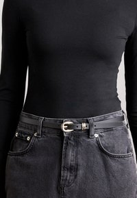 Cintura in pelle nera con fibbia dorata, indossata sopra una blusa nera a maniche lunghe e una gonna in denim grigio scuro, che mostra un design elegante e semplice.
