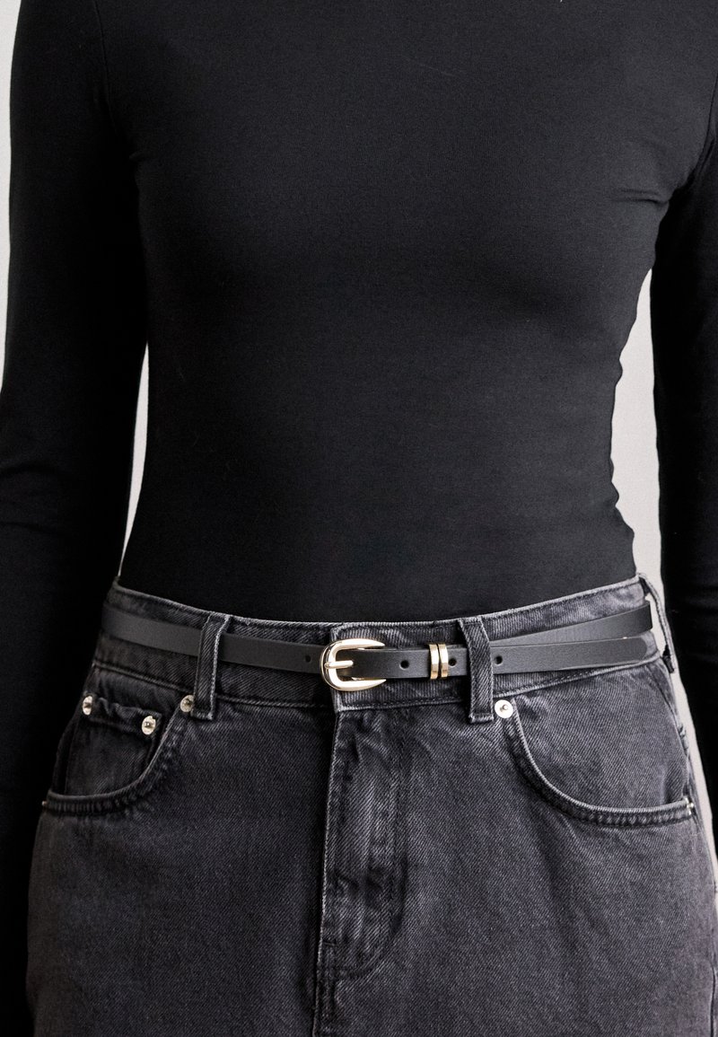 Cintura in pelle nera con fibbia dorata, indossata sopra una blusa nera a maniche lunghe e una gonna in denim grigio scuro, che mostra un design elegante e semplice.