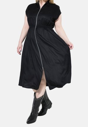 Femme portant une robe mi-longue noire avec fermeture éclair devant et taille cintrée, associée à des bottines noires cloutées, debout devant un fond blanc.