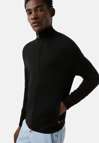 Černý svetr na zip s vysokým límcem. Má žebrované manžety, boční kapsy a hladkou texturu. Minimalistický design bez vzorů.