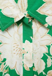 Korte mouw shirt met een groene achtergrond en grote beige bloemenpatronen, een puntige kraag en zichtbare stikdetails langs de naden.