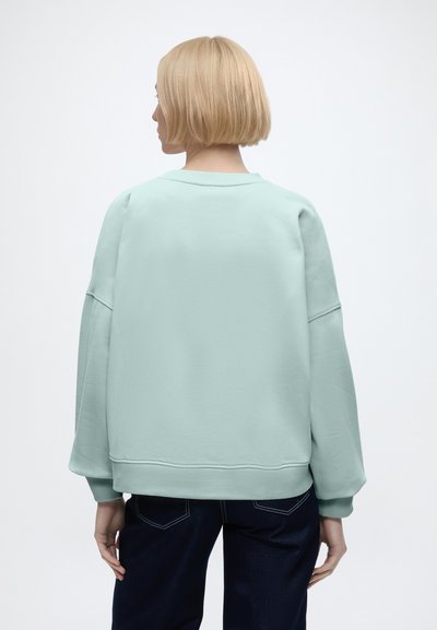 Personne aux cheveux blonds courts portant un sweat-shirt bleu clair et un pantalon foncé, tournée dos à la caméra sur un fond uni.