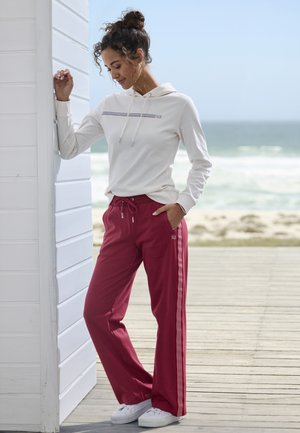 Helle beige Kapuzen-Sweatshirt mit horizontal gestreiftem Design, kombiniert mit roten tapered Jogginghosen, die weiße Seitestreifen und einen Kordelzug haben. Weiße Sneakers.