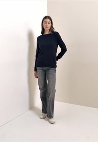 Marineblauer Strickpullover mit Rundhalsausschnitt, kombiniert mit grauen Jeans mit geradem Bein und weißen Turnschuhen vor neutralem Hintergrund.