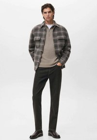 Giovane uomo che indossa una giacca a quadri grigia con bottoni sopra un maglione beige a mezza zip, pantaloni neri e stivaletti in pelle scura, in piedi con le mani nelle tasche.