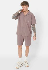 Helles mauve Kurzarmhemd mit einer Tasche, kombiniert mit passenden Shorts, beide aus weichem Stoff gefertigt. Tragen mit creamfarbenen Sneakers und weißen Socken.