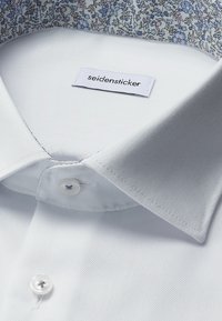 Close-up van de kraag van een wit overhemd met een bloemenpatroonvoering en een label van het merk "Seidensticker" aan de binnenkant van de kraag.