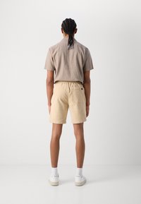 Chemise courte à manches, marron clair, short en velours côtelé beige avec taille élastique et baskets blanches. Texture lisse et palette de couleurs neutres.