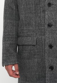 Gray wool coat featuring a check pattern, with black buttons and a flap pocket on the front. The fabric has a textured finish.