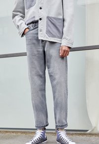 Szara kurtka na guziki z kontrastowymi szarymi kieszeniami, w połączeniu z jasnoszarymi dżinsami z podwiniętymi mankietami i niebieskimi wysokimi trampkami.