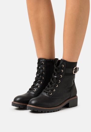 Bottes noires en cuir à chevilles avec un laçage à l'avant, une fermeture éclair sur le côté et une sangle avec une boucle métallique. Semelle texturée avec un léger talon carré.