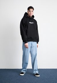 Black hooded sweatshirt with white logo, paired with light blue tracksuit pants. Black sneakers with white soles, standing on a blue carpet.