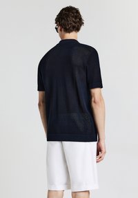 Mann trägt ein dunkelblaues kurzärmliges Strickshirt und weiße knielange Shorts, von hinten vor einem schlichten Hintergrund betrachtet.