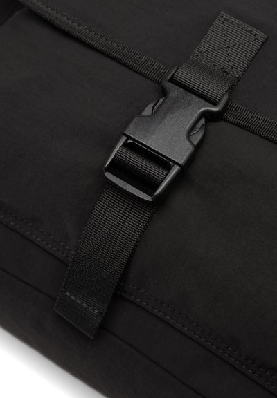 Sac en tissu noir avec une boucle en plastique durable et une sangle en toile, présentant des coutures visibles et une texture lisse.