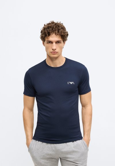 Tumši zils kokvilnas t-krekls ar īsām piedurknēm, pieguļošs dizains un neliels logotips kreisajā krūtīs. Kombinēts ar pelēkām biksēm.