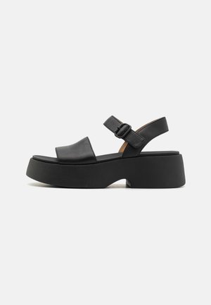 Sandali platform in pelle nera con una larga fascia e fibbia regolabile alla caviglia, caratterizzati da una suola in gomma spessa e una texture liscia.