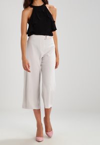 Schwarzes Neckholder-Oberteil mit Rüschen, kombiniert mit weißen, weit geschnittenen Hosen und hellrosanen High Heels. Glatter Stoff, strukturiertes Design und neutrale Farbtöne.