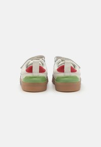 Sneakers med en kremfarget læroverdel, røde og grønne detaljer på hælen, og en kraftig gjennomskinnelig såle. Bakfra ser vi borrelåsremmer.